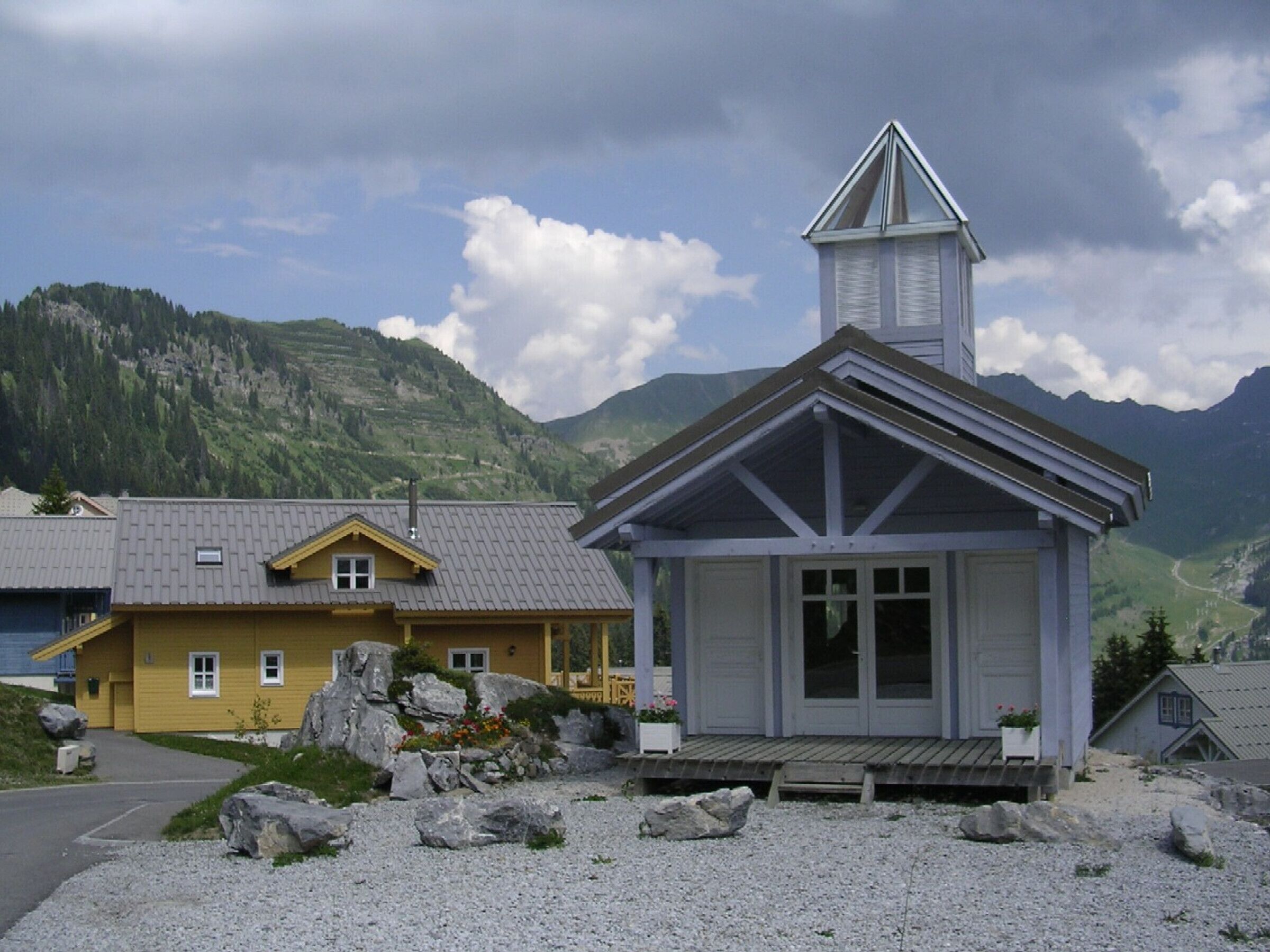 A small chapel with a bell tower, surrounded by rocky terrain and mountains in the background.