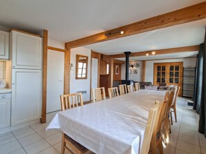 A dining area with a long table, wooden chairs, and a kitchen in the background.