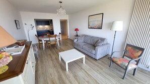 Ein modernes Wohnzimmer mit grauem Sofa, einem weißen Couchtisch und einem Essbereich mit Tisch und Stühlen. Im Hintergrund ist eine Küche zu sehen.
