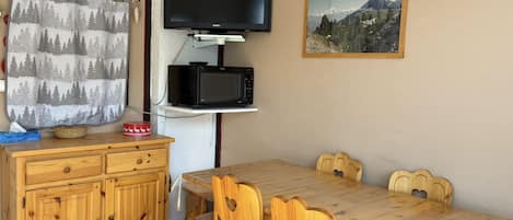 A dining area with a wooden table and chairs, a television mounted on a shelf, and a mountain landscape painting on the wall.