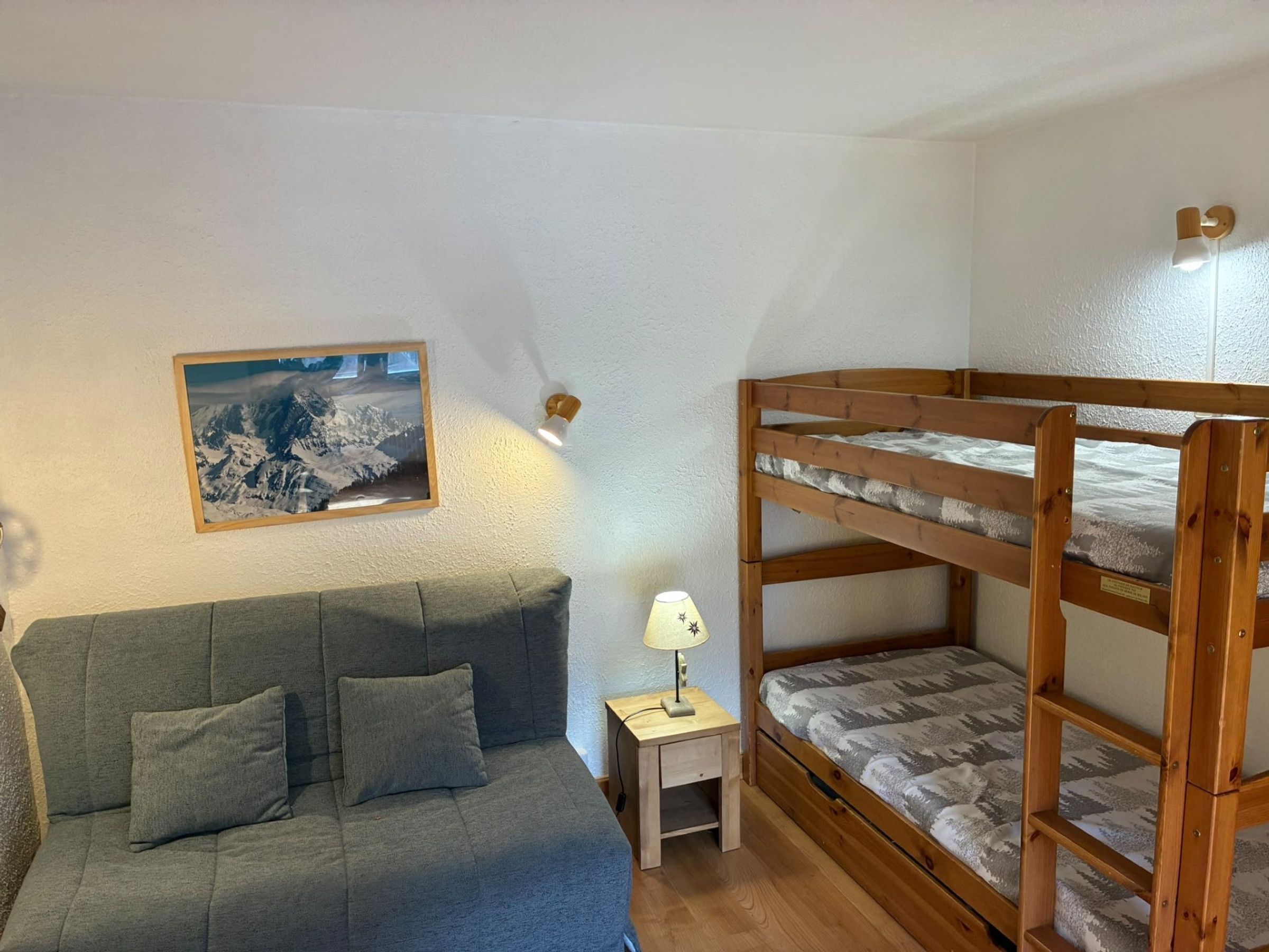 A room with a bunk bed, a sofa, a small bedside table, a lamp, and a framed picture on the wall.