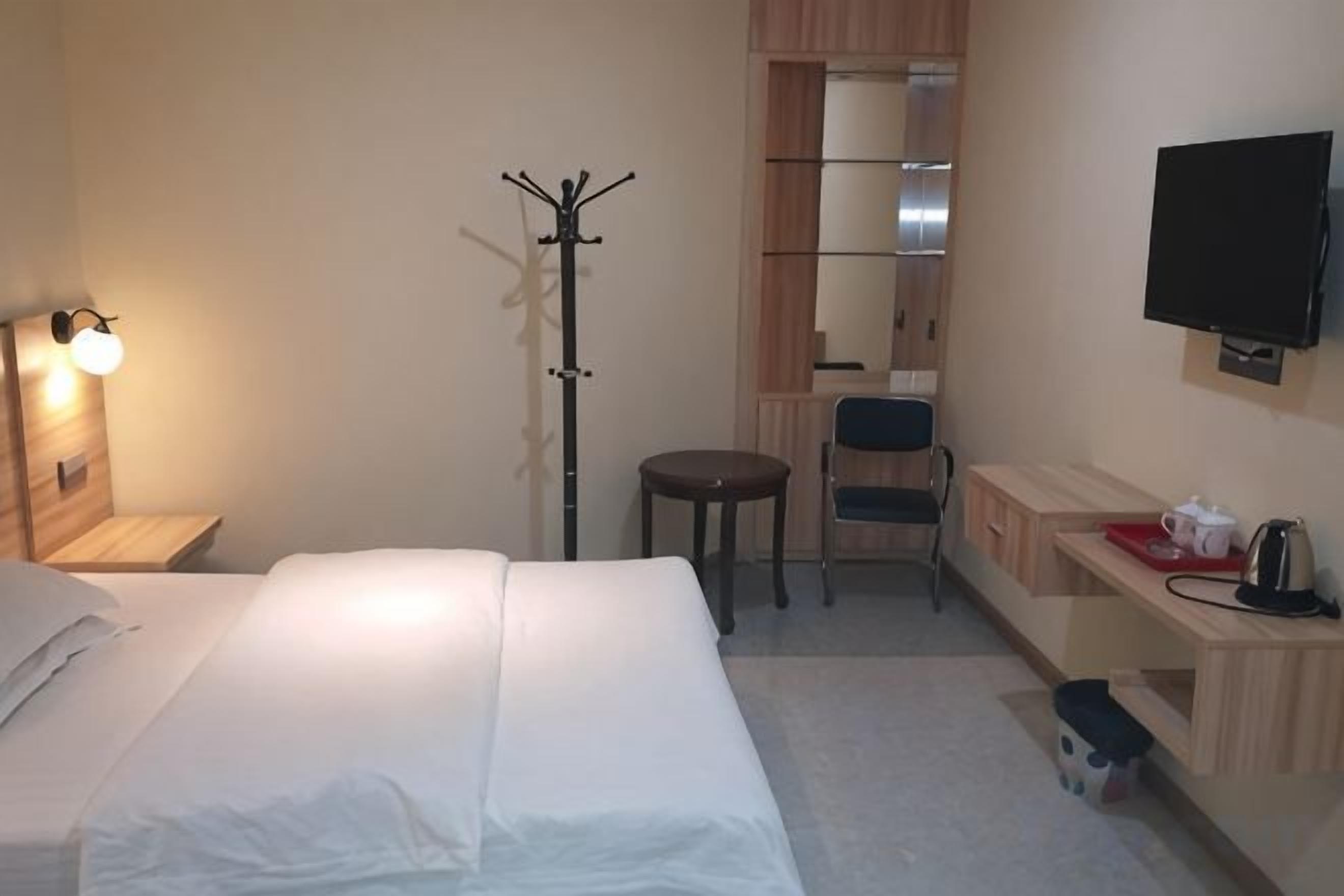Habitación de hotel con cama, escritorio, silla, perchero, televisión y una mesita con hervidor.