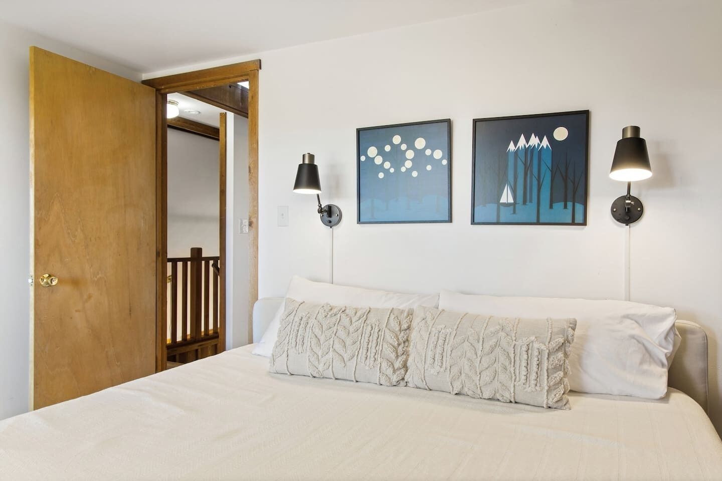 A bedroom with a bed, two wall-mounted lamps, and two framed artworks.