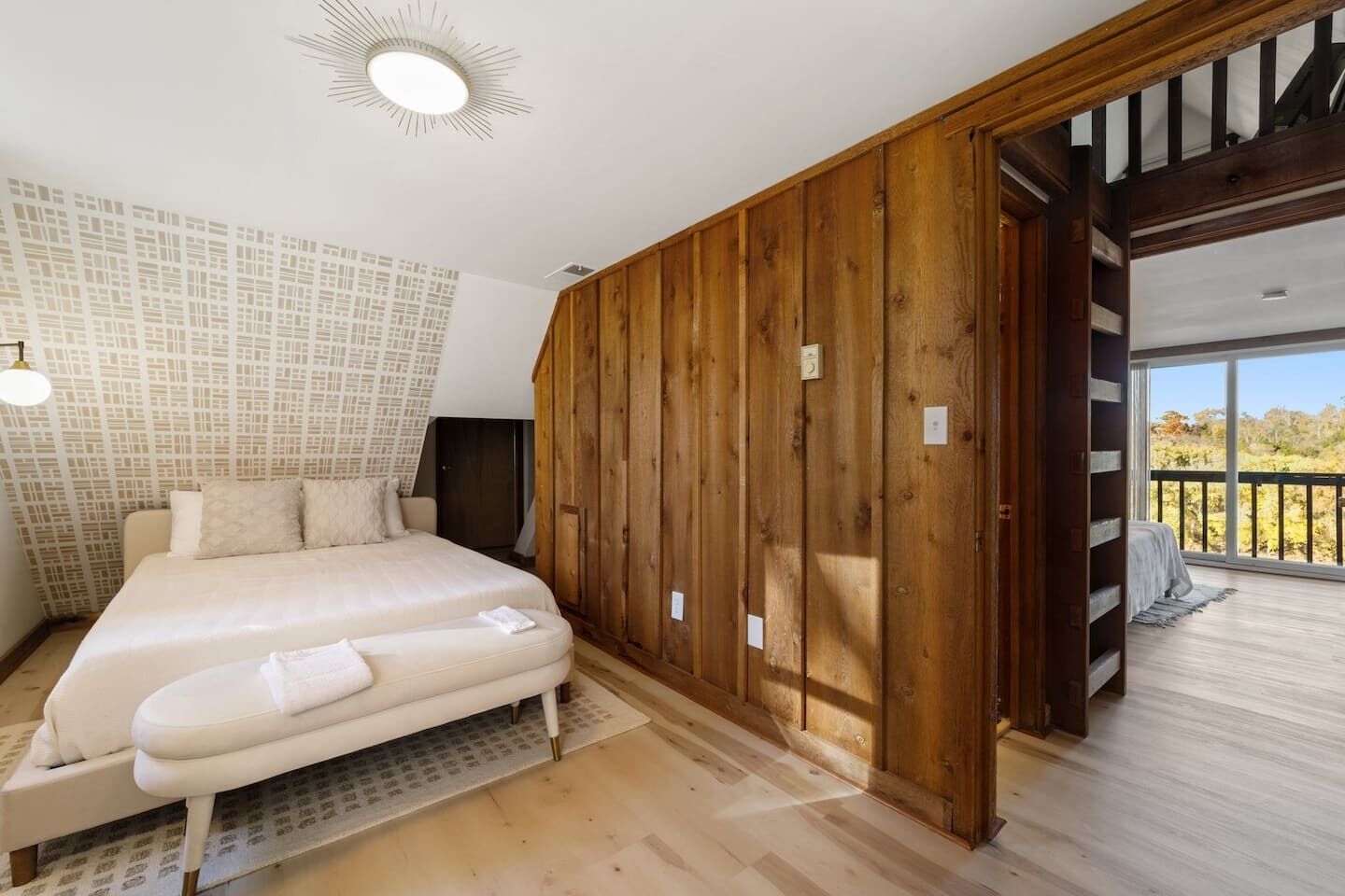 A bedroom with a large bed, wooden paneling, a ceiling light, and a balcony with a view.