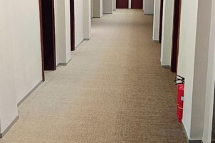 Pasillo alfombrado con puertas a ambos lados, que conducen a habitaciones. Un extintor está montado en la pared.