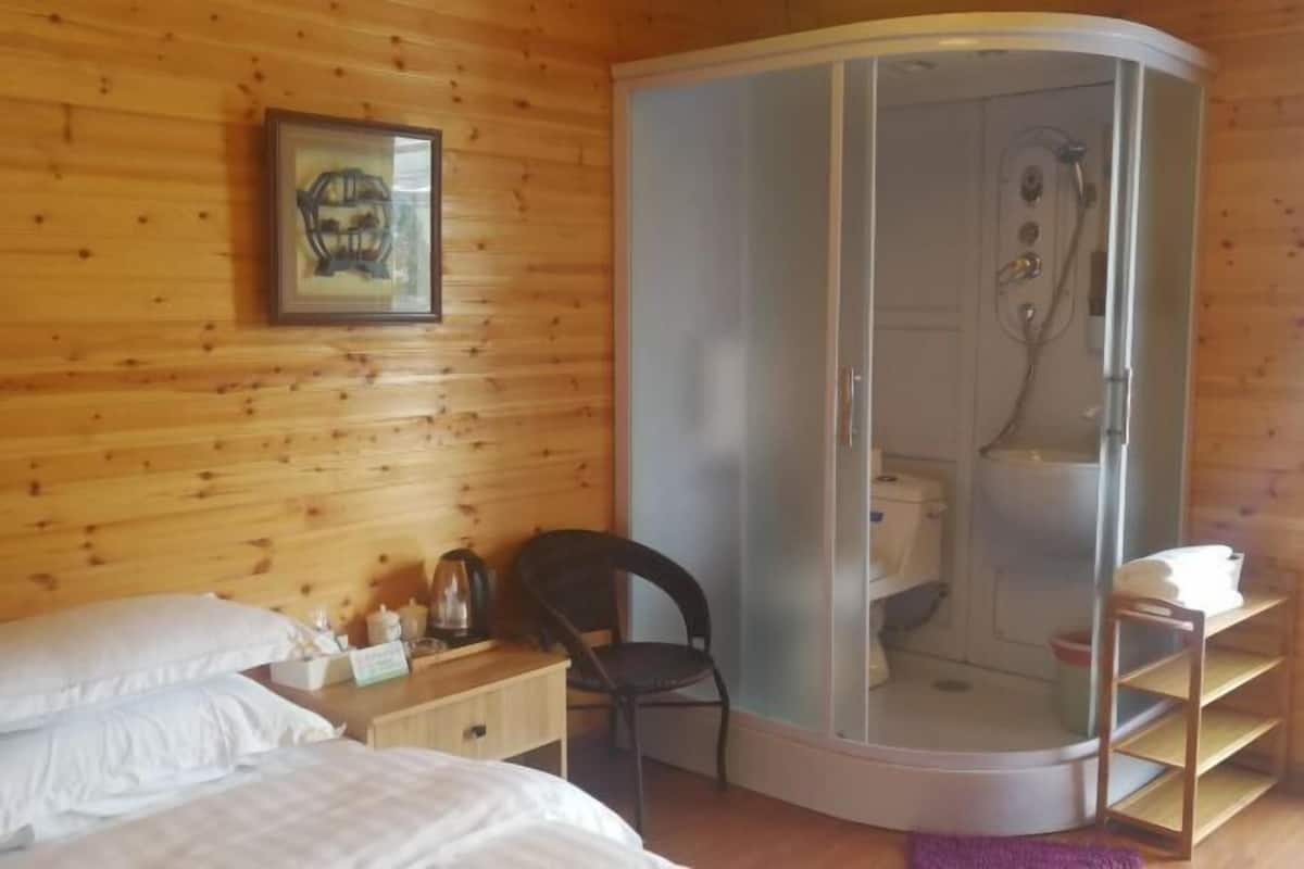 A room with wooden walls, a bed with white bedding, a chair, a shower enclosure, and a shelf with towels.