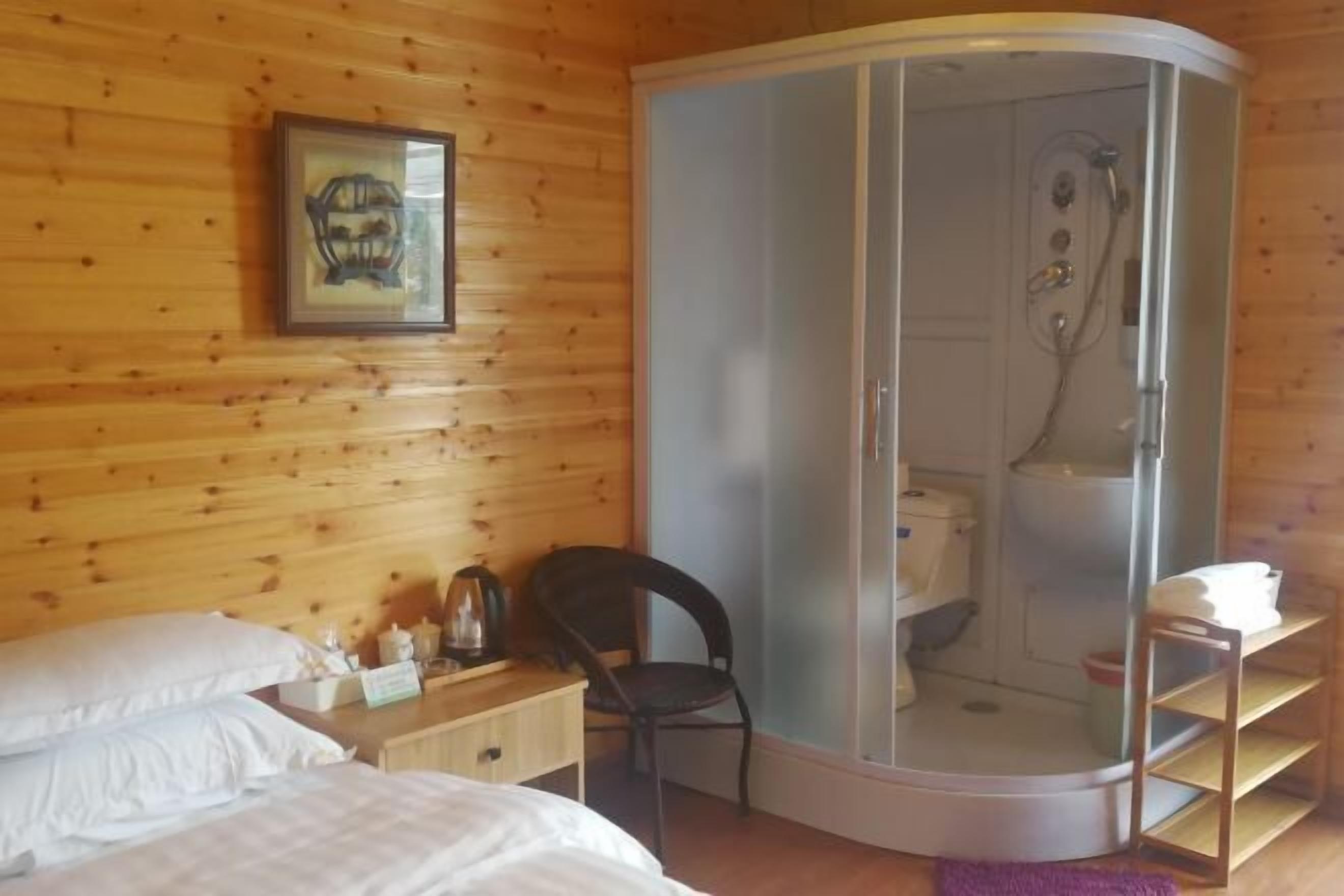 Une pièce aux murs en bois, un lit avec du linge de lit blanc, une chaise, une petite table avec des articles de toilette et un espace douche.