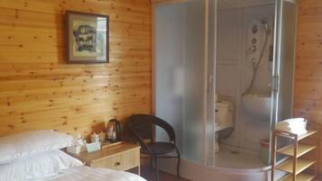 Une pièce aux murs en bois, un lit avec du linge de lit blanc, une chaise, une petite table avec des articles de toilette et un espace douche.