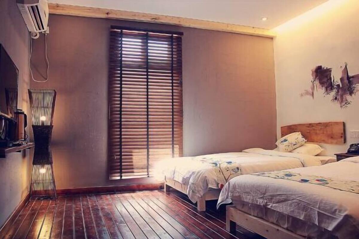 Ein Zimmer mit zwei Betten, Holzboden, Fenster mit Jalousien, Wand-Klimaanlage und einem Nachttisch mit Lampe.