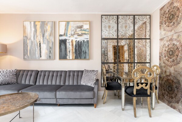 Ein modernes Wohnzimmer mit grauem Sofa, rundem Couchtisch und einem Essbereich mit Holzstuhl und Glastisch. An der Wand hängen abstrakte Gemälde und ein dekorativer Spiegel.