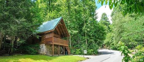 A rustic cabin with a stone foundation and a wooden deck, surrounded by dense forest.