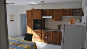A compact kitchen with wooden cabinets, a refrigerator, and a dining table with a colorful tablecloth. - Havre de Paix (Fontenoy-le-Château)
