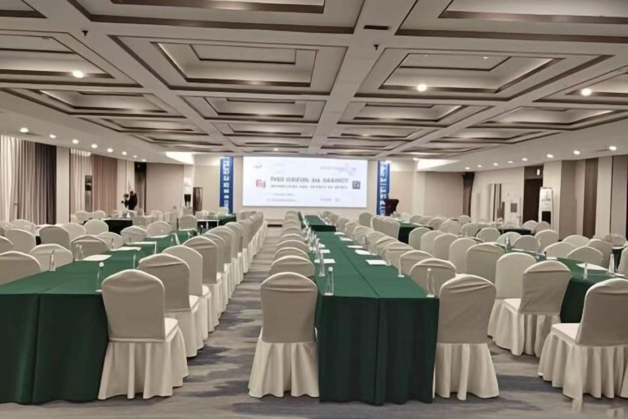 A conference room with rows of tables and chairs, a projector screen, and a presentation on the wall.