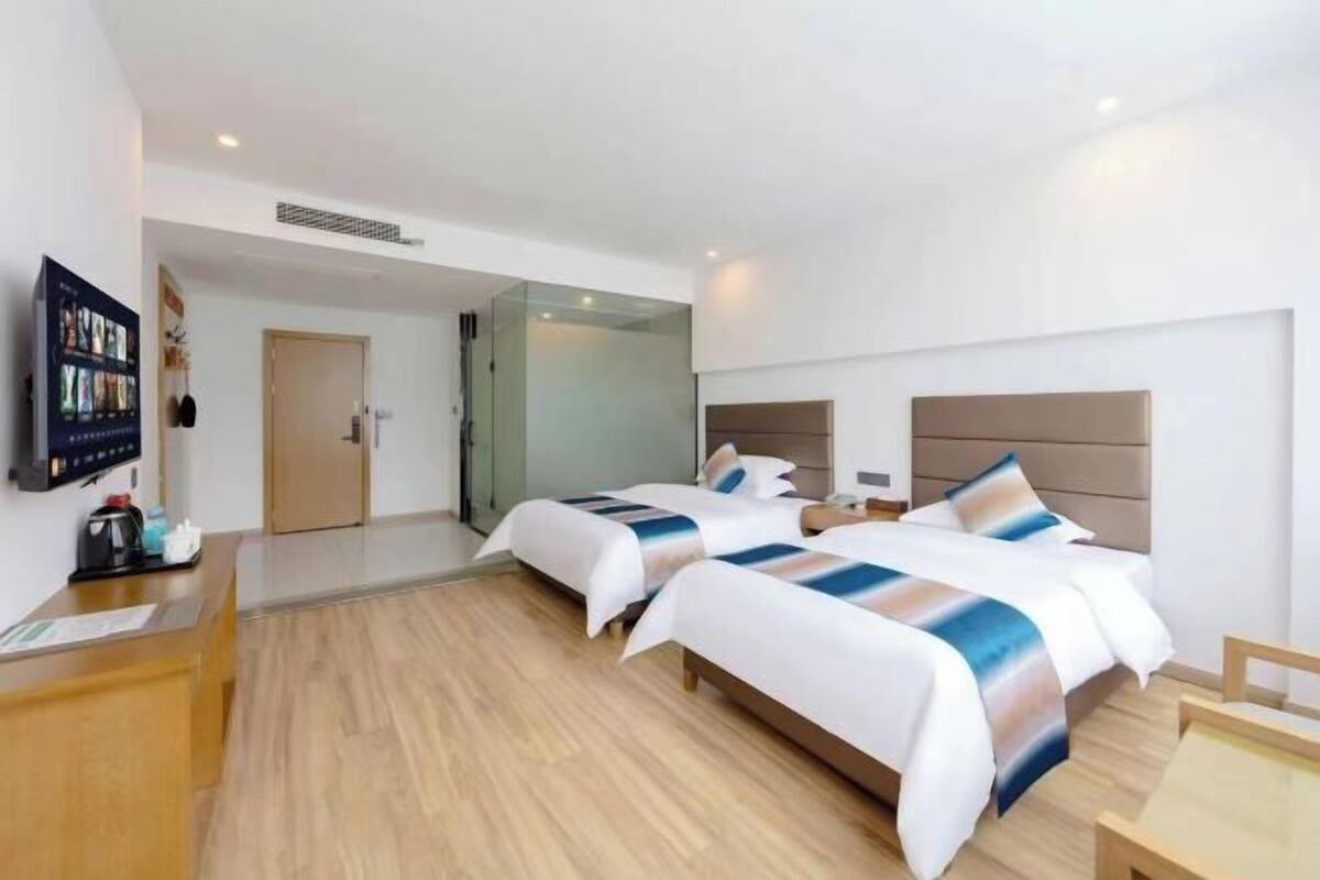 Quarto de hotel com duas camas, uma escrivaninha, televisão e um painel de vidro.