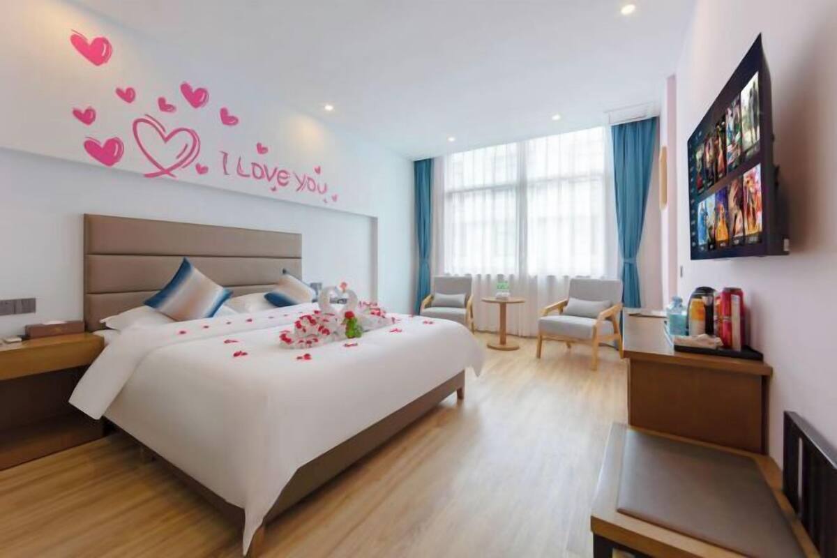 Quarto de hotel com cama, escrivaninha, cadeira, televisão e uma parede decorada com um coração.
