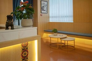 Zona de recepción de un hotel con mostrador, una planta en maceta, una mesita y un ventanal con cortinas.