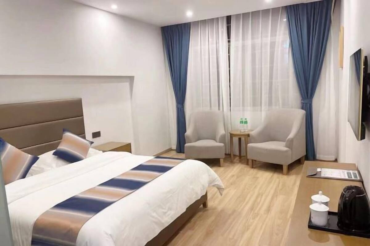 Quarto de hotel com cama, duas poltronas, uma mesinha com chaleira e televisão na parede.