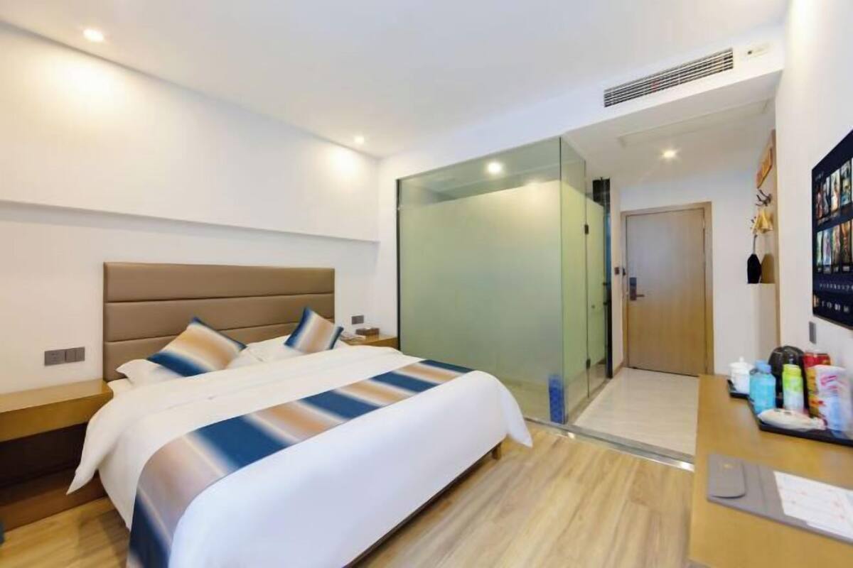 Quarto de hotel moderno com uma cama grande, uma escrivaninha com artigos de higiene e uma divisória de vidro.