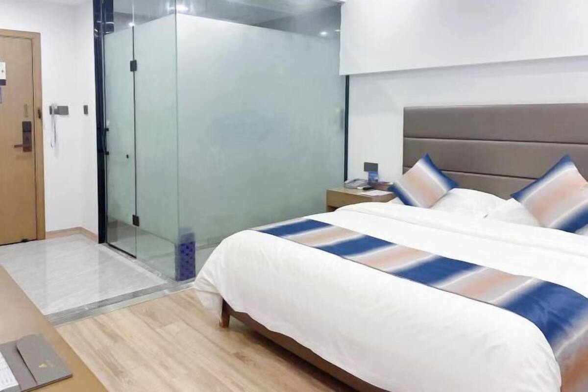 Quarto de hotel com cama, mesa de cabeceira, chuveiro em cabine de vidro e porta de madeira.