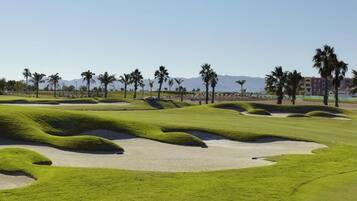 Um campo de golfe com bunkers de areia, palmeiras e montanhas ao longe.