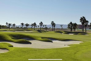Um campo de golfe com bunkers de areia, palmeiras e montanhas ao longe.