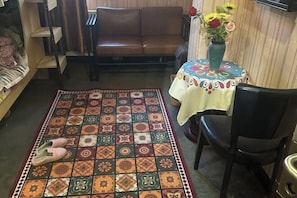 Un salon avec un tapis à motifs, un canapé en cuir marron, une table ronde recouverte d’une nappe colorée et un vase rempli de fleurs.