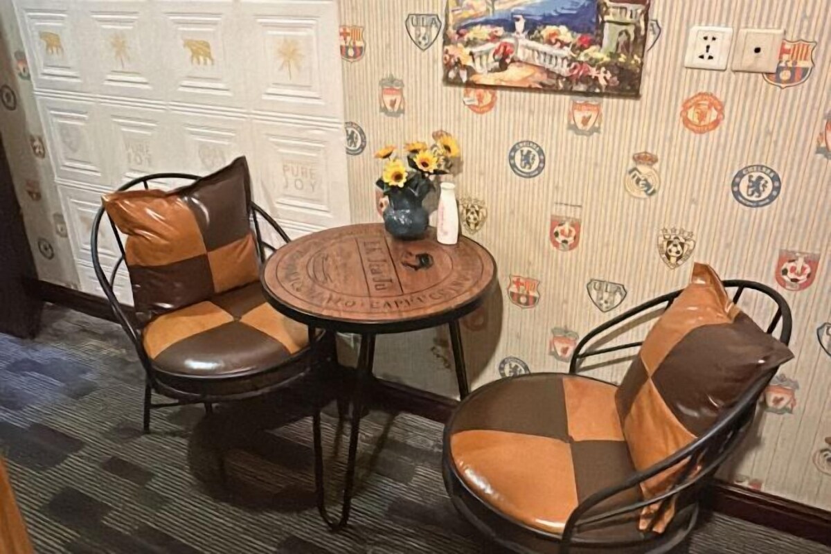 A room with two metal chairs with brown cushions, a small round table with a vase of sunflowers, and a wall decorated with various sports team logos.