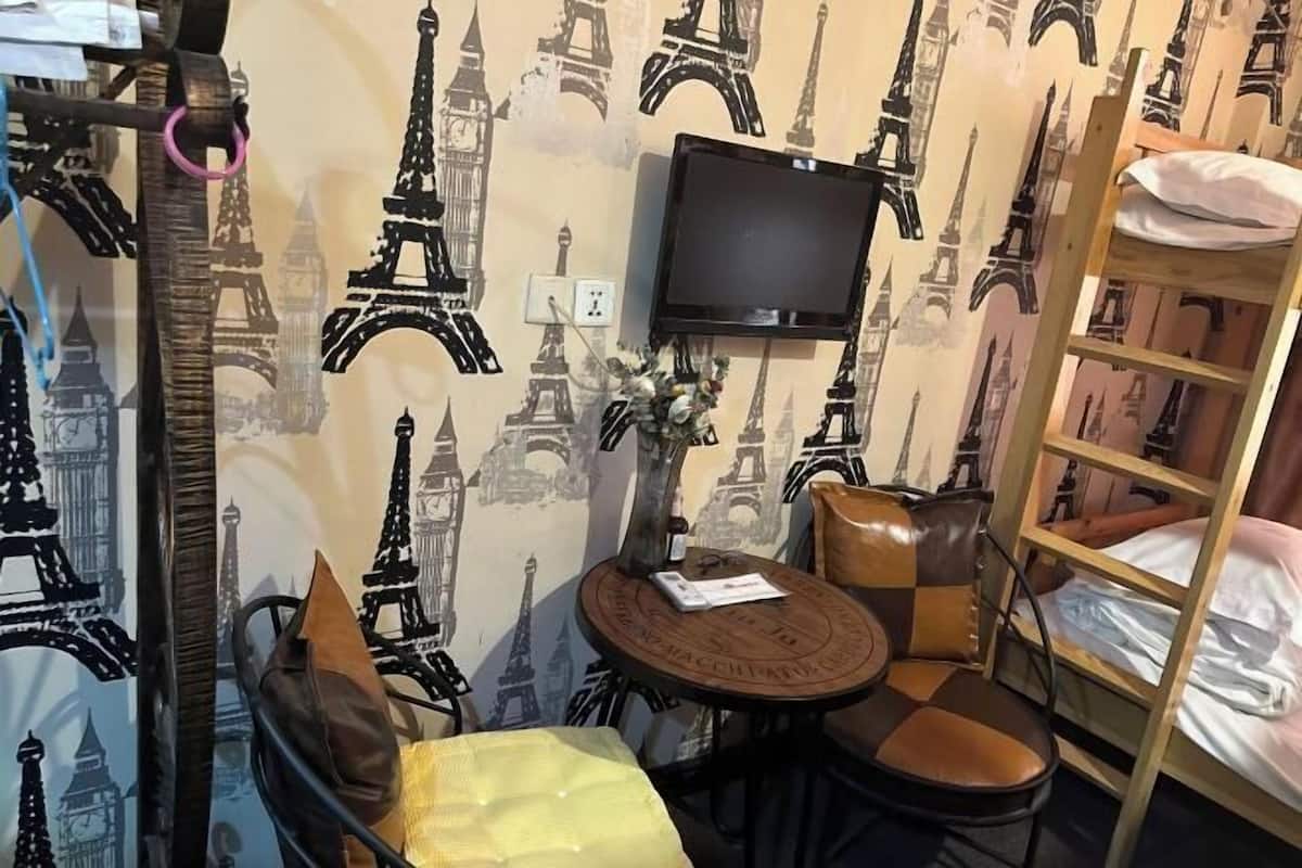 Um quarto com cama, televisão presa à parede, mesa redonda com cadeiras e uma estante de livros.