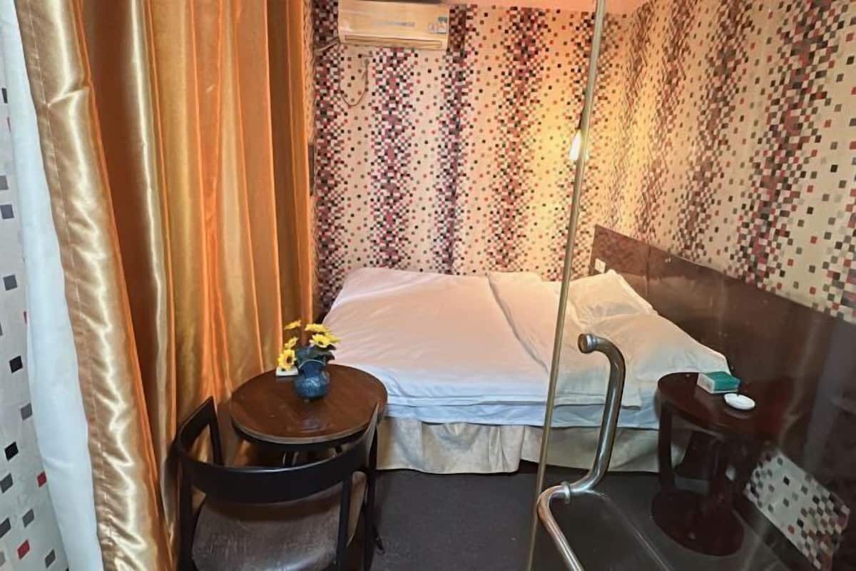 Quarto pequeno e aconchegante com cama, uma mesinha com um vaso de flores, uma cadeira e uma mesinha de cabeceira com um porta-lenços.
