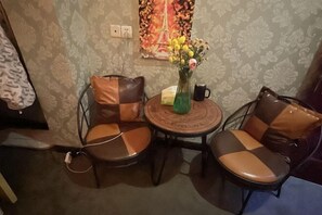 Um quarto pequeno com duas cadeiras, uma mesa, um vaso com flores e um quadro na parede.