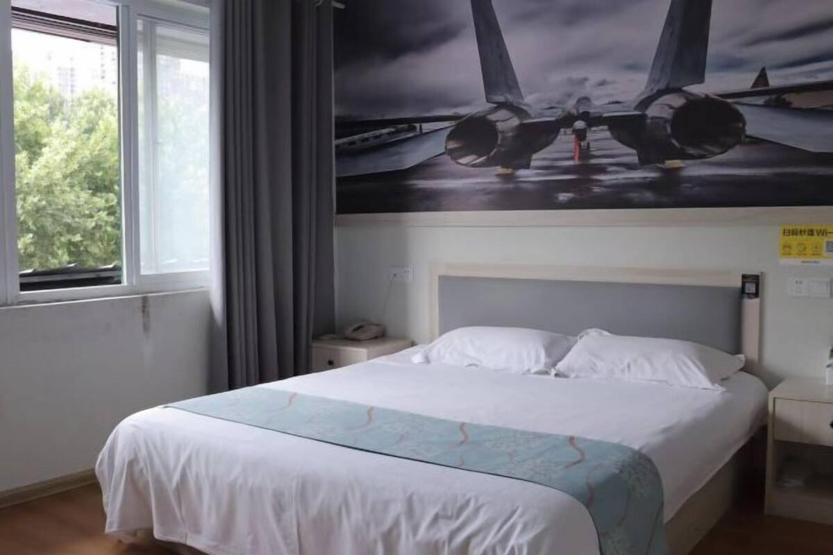 Habitación de hotel con cama, mesita de noche, ventana con cortinas e imagen grande de un avión en la pared.