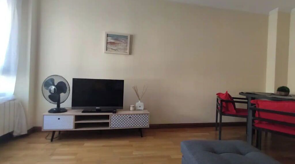 Un salon avec un téléviseur posé sur un meuble, un ventilateur, une petite table avec une bougie et un fauteuil rouge.