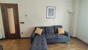 Un salón con un sofá seccional azul oscuro, dos cojines color beige, una obra de arte enmarcada en la pared, una lámpara de pie alta y suelo de madera.