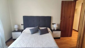 Una cama bien hecha con cabecera gris, dos mesitas de noche con lámparas y un armario de madera.