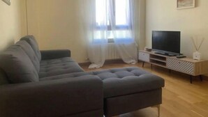 Una sala de estar con un sofá gris, un televisor de pantalla plana sobre un pedestal de madera, una lámpara de pie y un cuadro en la pared.