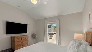 Un dormitorio con cama, cómoda de madera, televisor montado en la pared y ventana con cortinas.