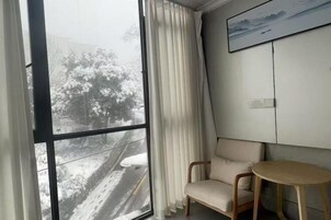 Um quarto com uma janela grande, uma cadeira, uma mesinha e um quadro na parede.