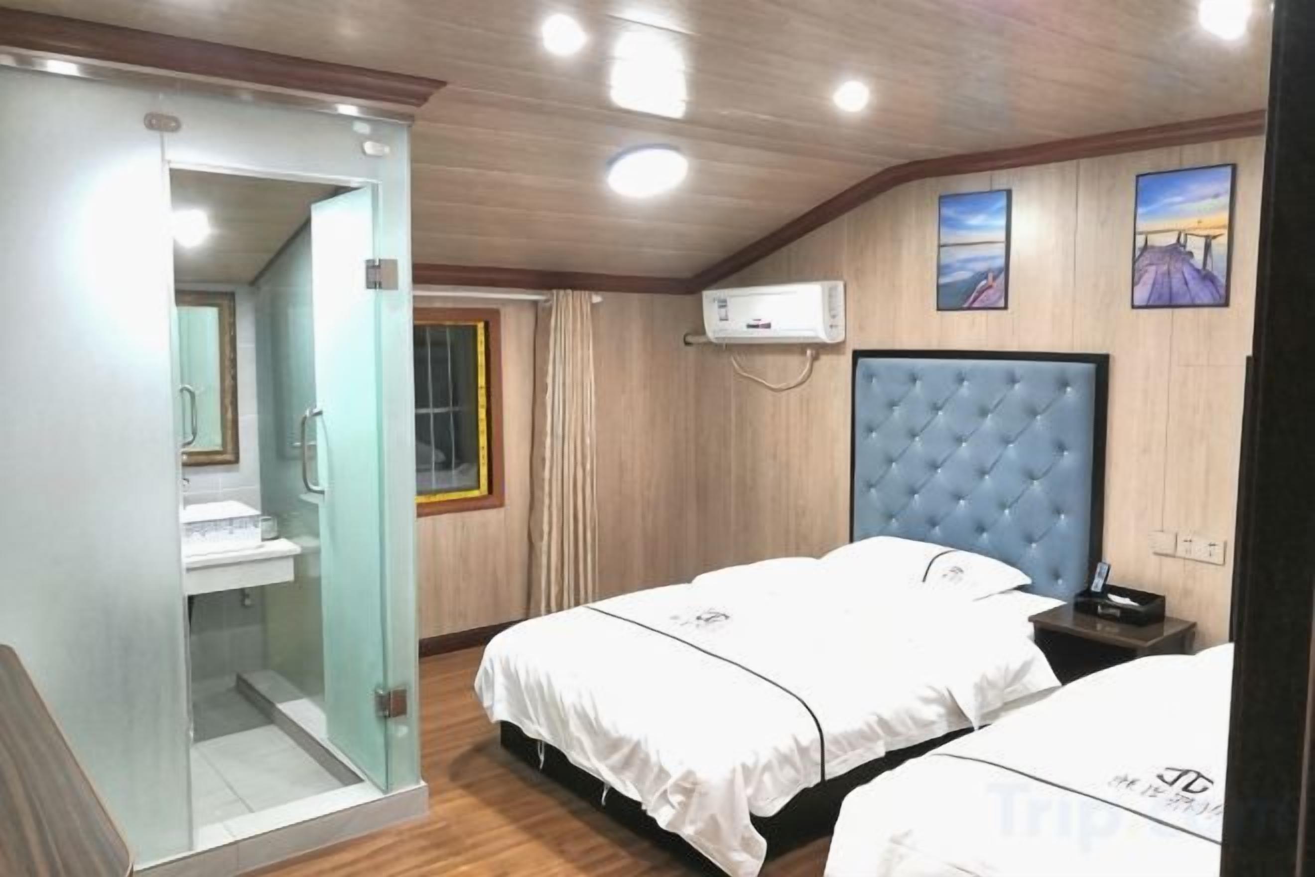 A hotel room with a glass-enclosed shower, a bed with a blue headboard, and a wall-mounted air conditioning unit.