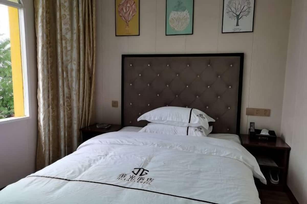 Un lit bien fait, avec une couette blanche et des oreillers, une table de chevet en bois avec un téléphone, et une fenêtre avec des rideaux.