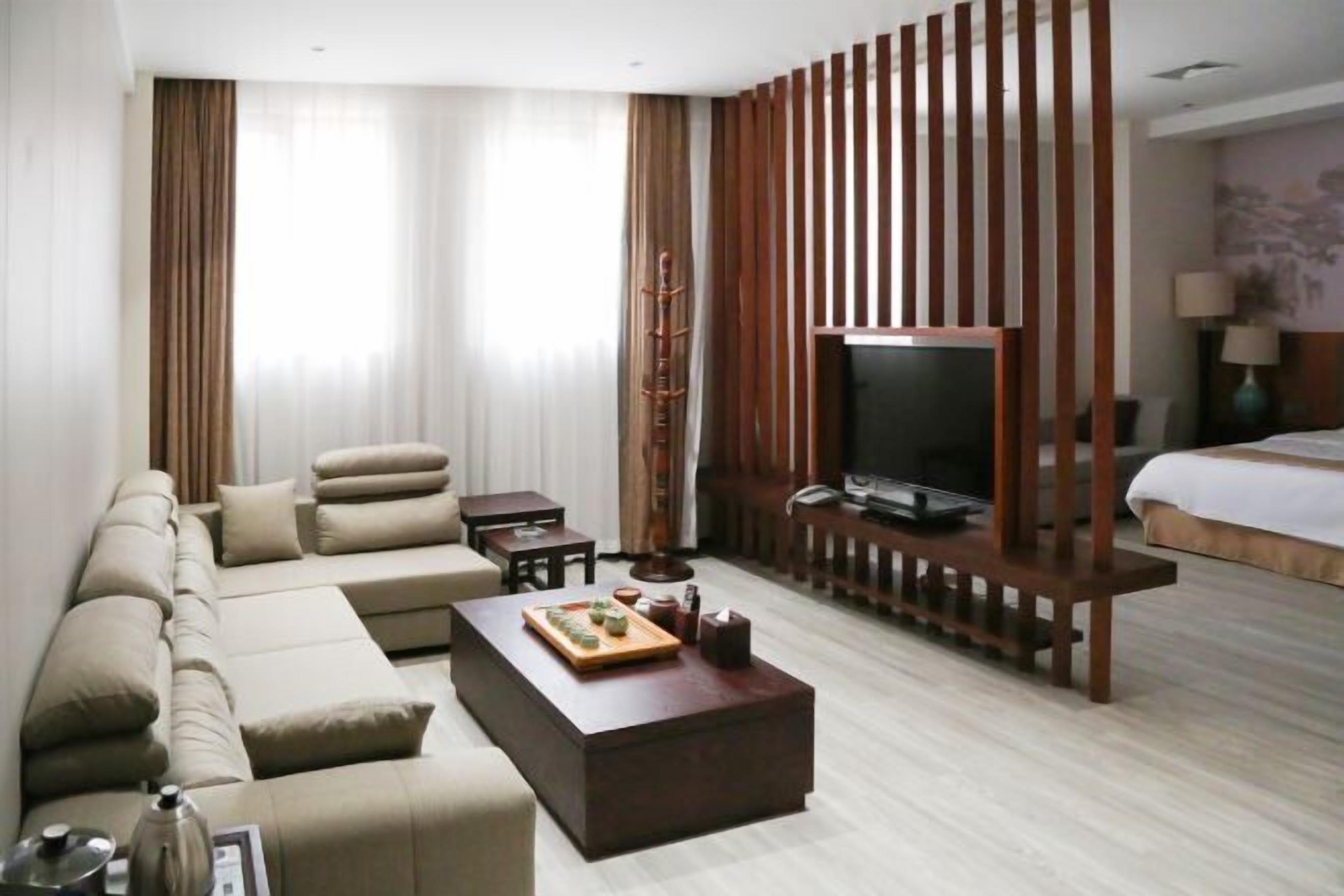A modern hotel room with a sectional sofa, a flat-screen TV, a wooden coffee table, and a bed with a headboard.