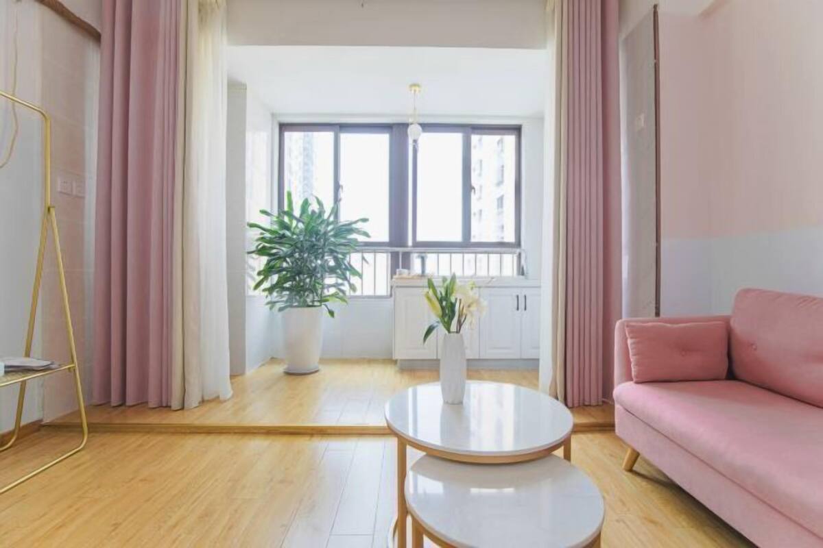 A modern living room with a pink sofa, two round tables, a potted plant, and a view of a balcony through large windows.