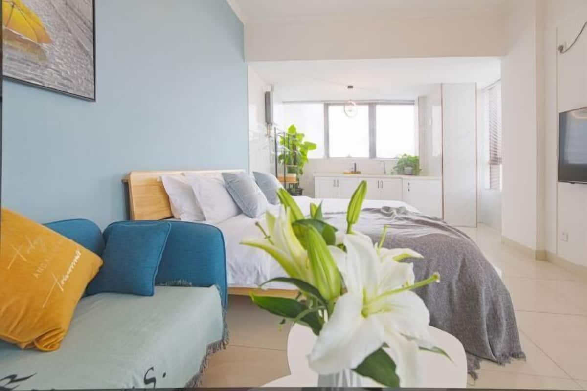 A modern bedroom with a bed, a sofa, a TV, and a kitchen in the background.