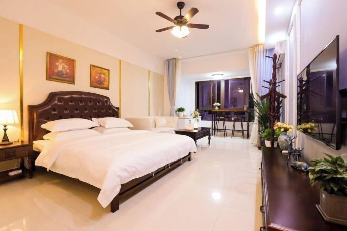 A hotel room with a large bed, a desk, a chair, a TV, and a ceiling fan.