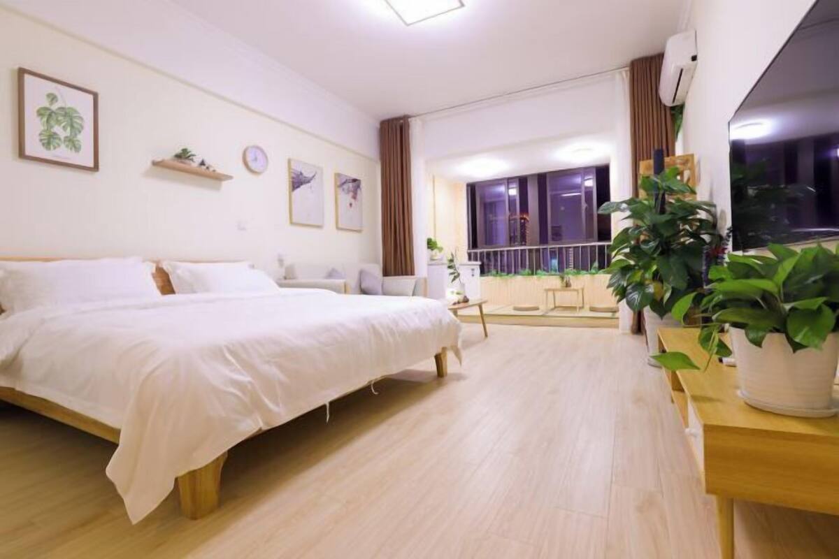 A modern hotel room with a large bed, a small desk, a balcony with plants, and a view of the city at night.