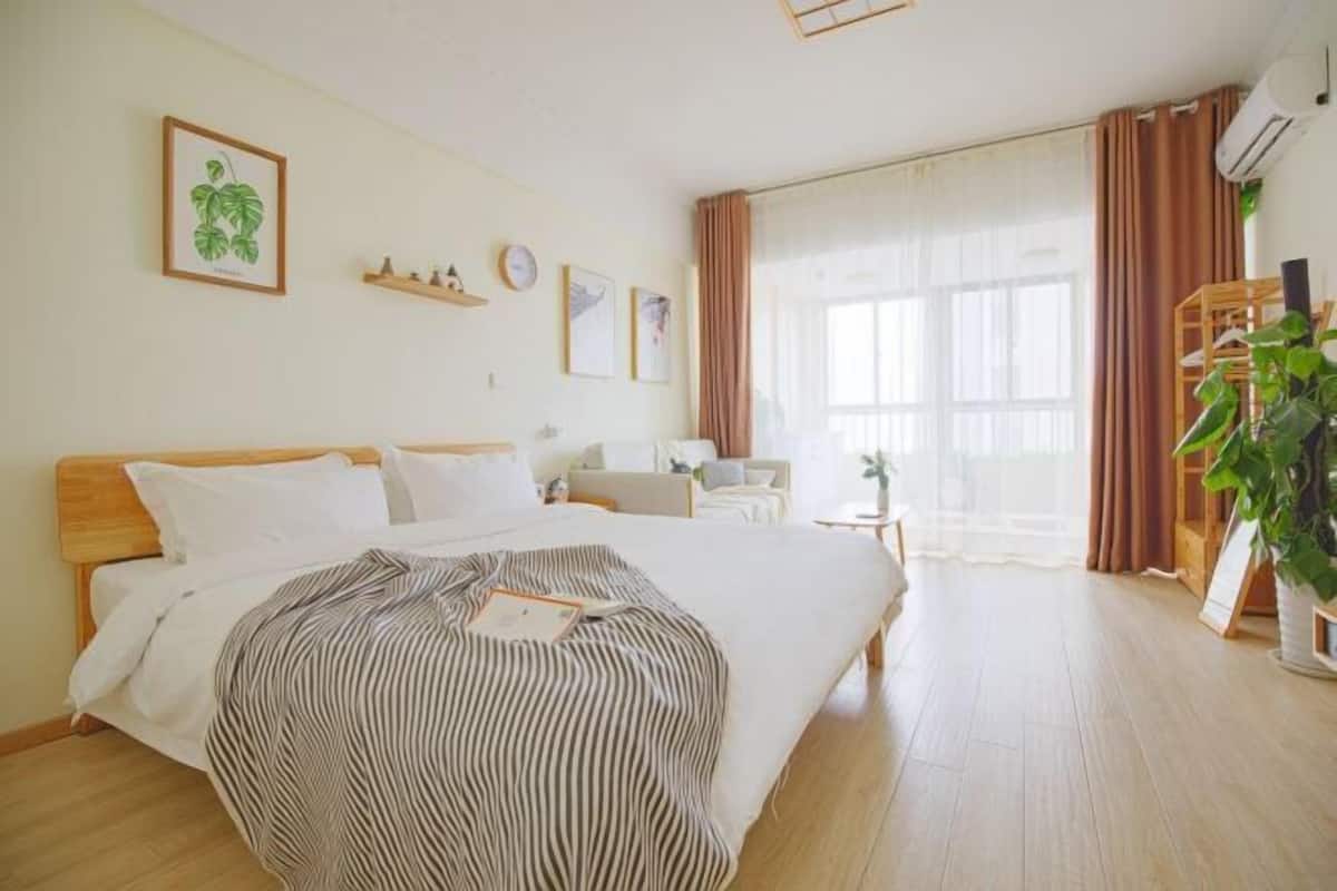 A bedroom with a bed, a bedside table, a chair, a window with curtains, and a potted plant.