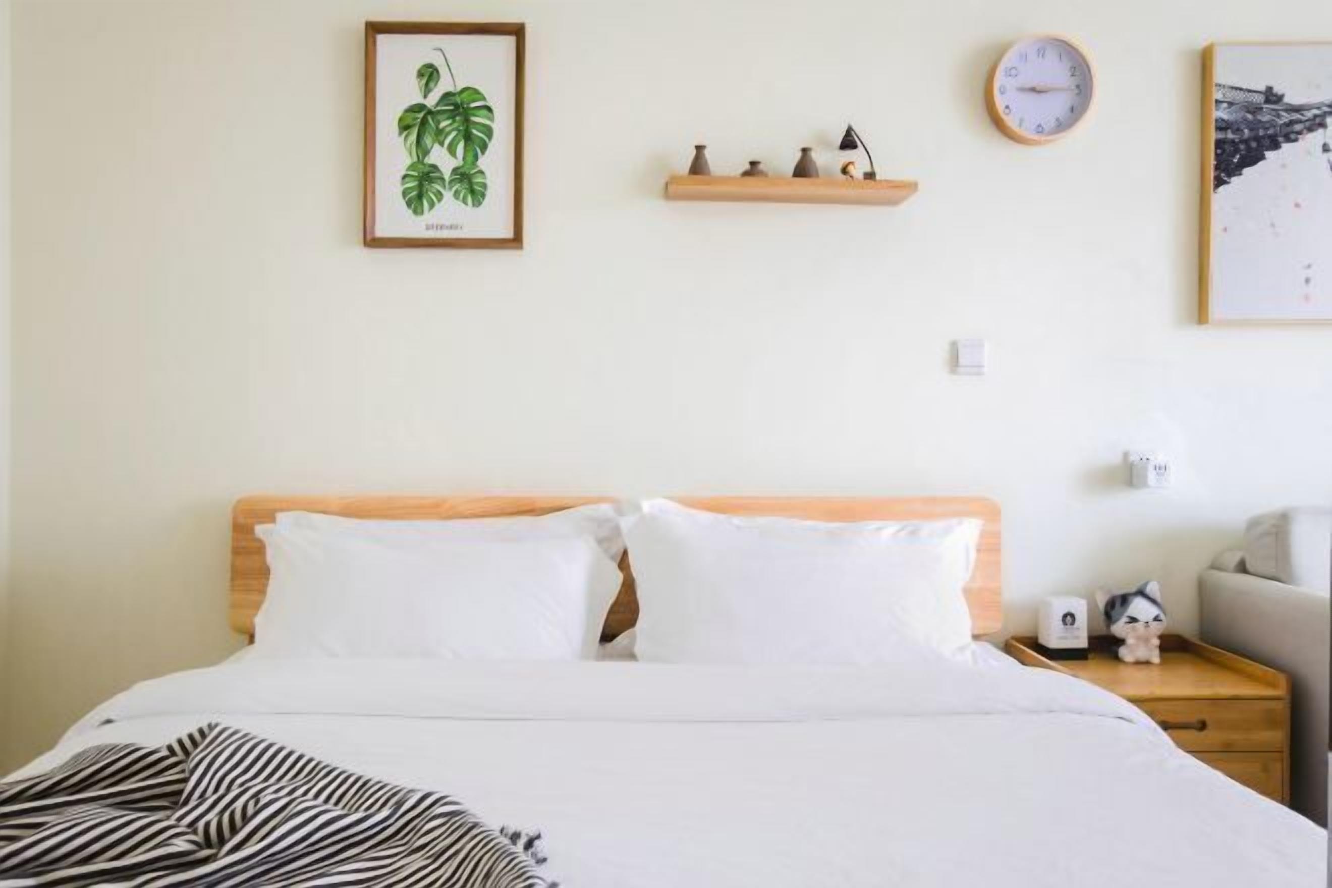 A neatly made bed with white bedding, a framed plant on the wall, a shelf with decorative items, a wall clock, and a small nightstand with a lamp and a figurine.