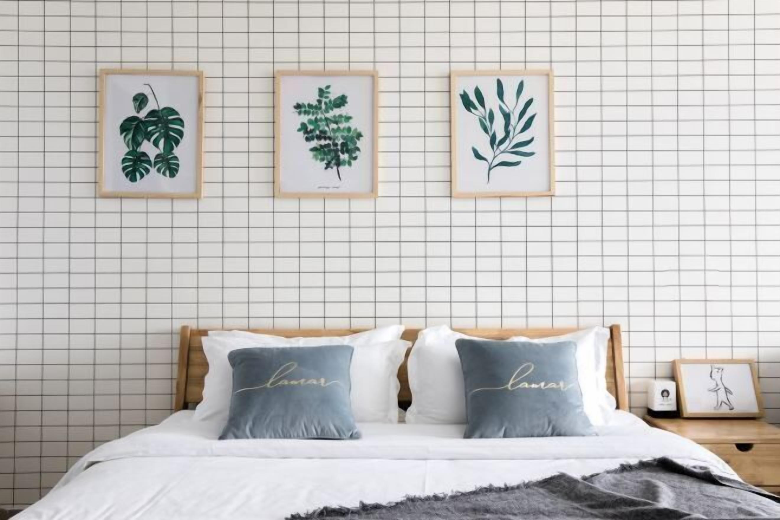 A neatly made bed with two pillows, a bedside table with a framed picture, and three framed botanical prints on the wall.