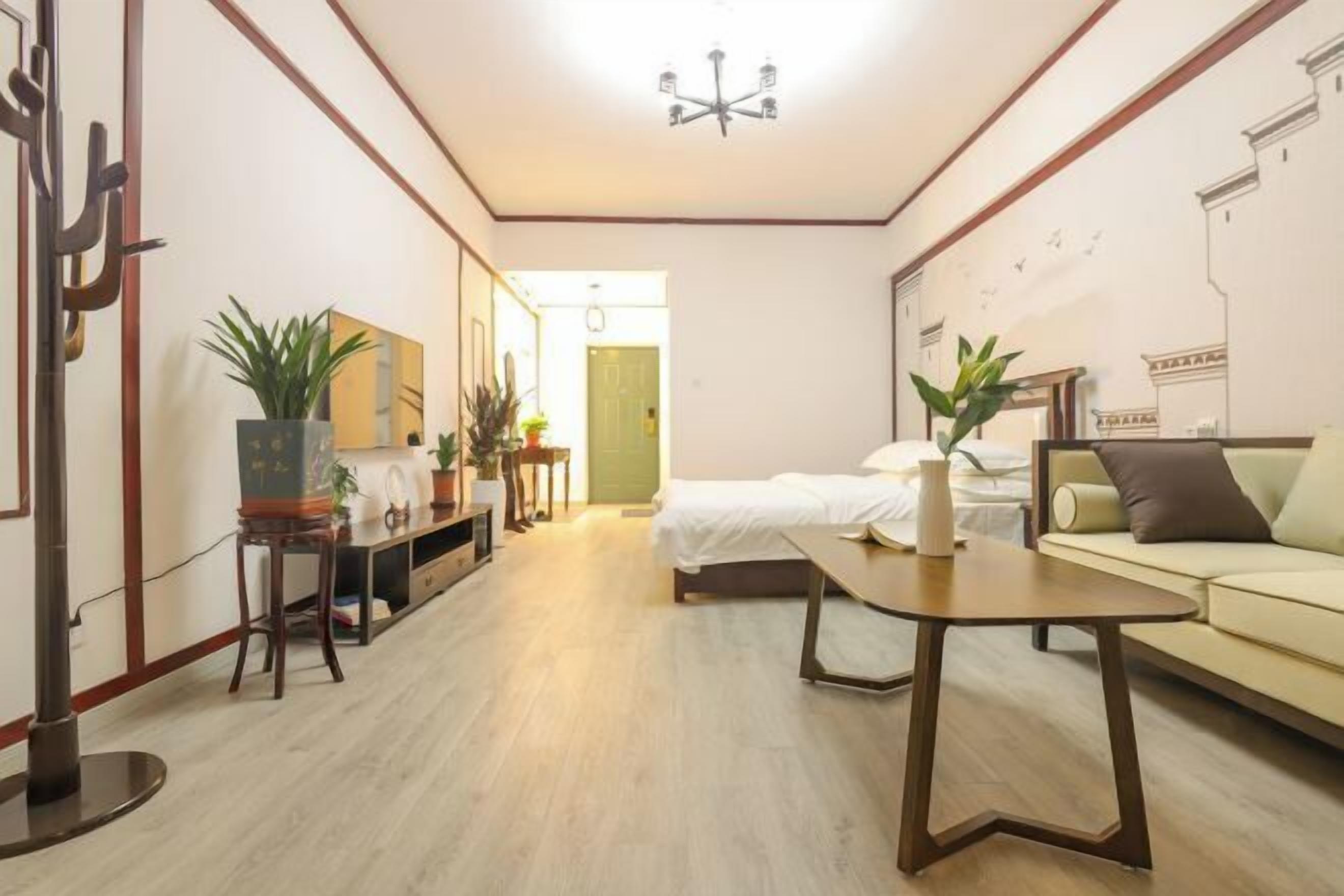A hotel room with a bed, a sofa, a small table, a desk, and a chair. There are potted plants and a mirror on the wall.