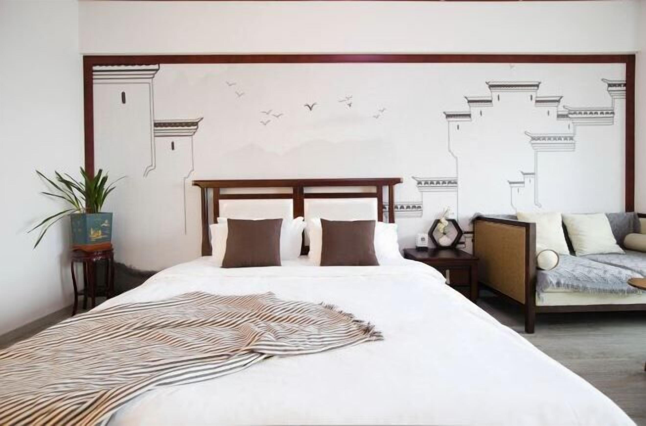 A bedroom with a large bed, a nightstand, a sofa, a wall mural, and a potted plant.