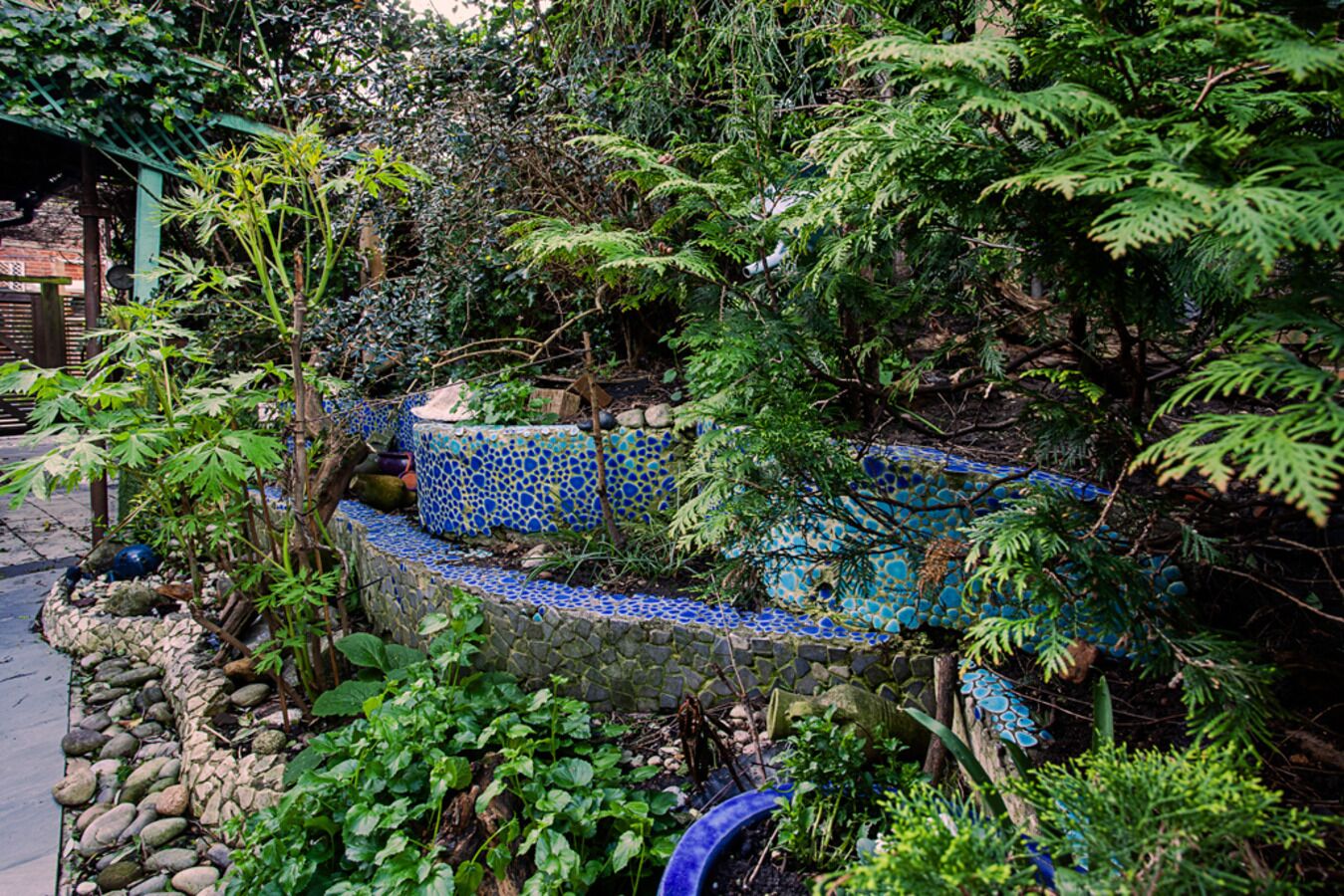 A garden with a mosaic-tiled bench surrounded by lush greenery and a stone wall.